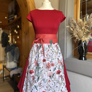 Schneiderpuppe mit kirschrotem Kleid, rotem Gürtel und weißer Blumenschürze vor Holzrahmen und Fenster